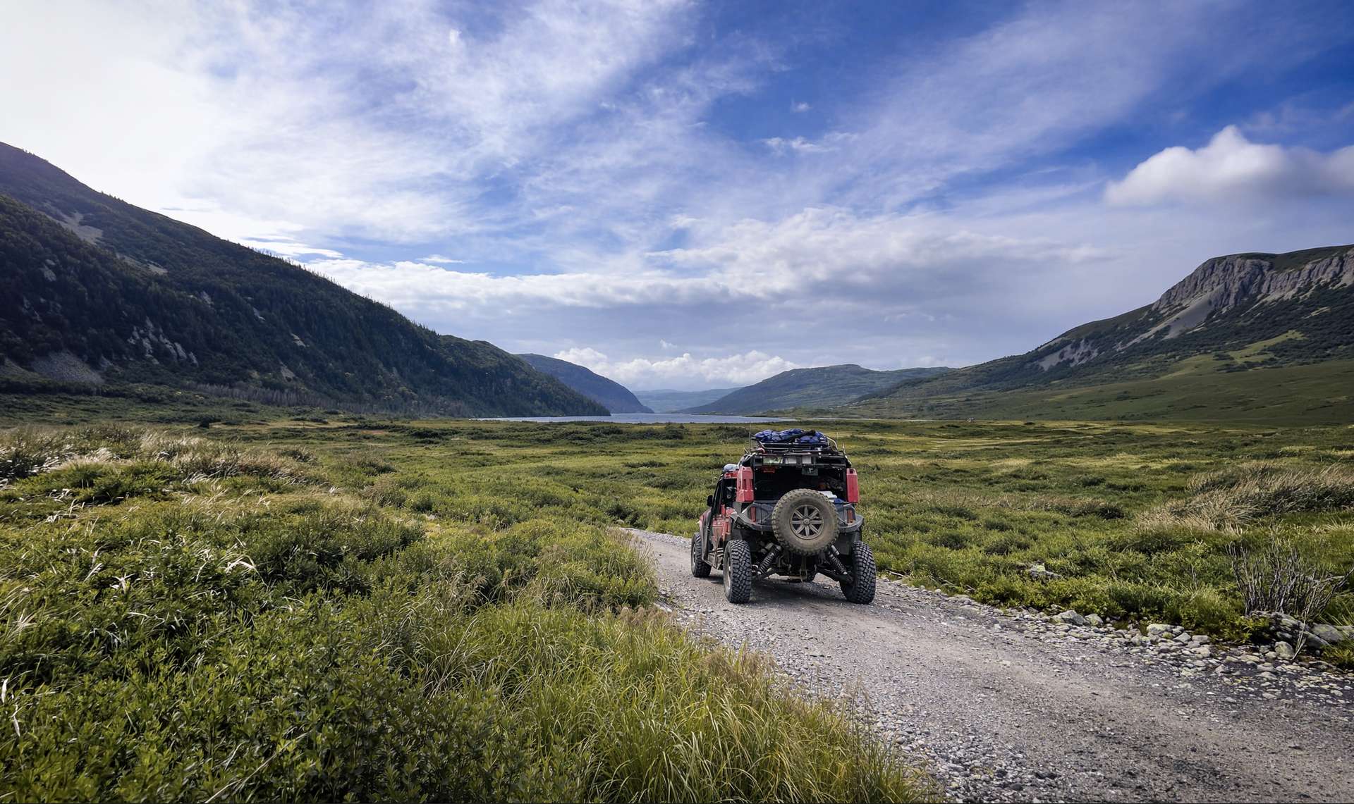 A red side-by-side on a gravel track in a wide green valley, with a lake and mountains ahead
