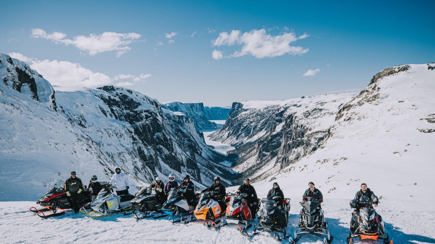 A line of riders and snowmobiles on a snowy ridge above a deep, frozen gorge in Newfoundland