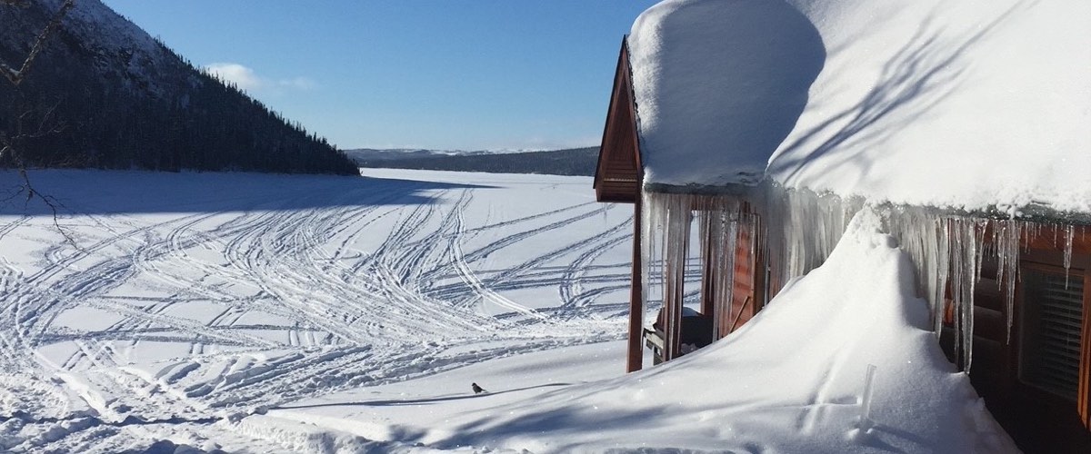 A log building under a heavy roof of snow beside a frozen lake, crisscrossed with snowmobile tracks