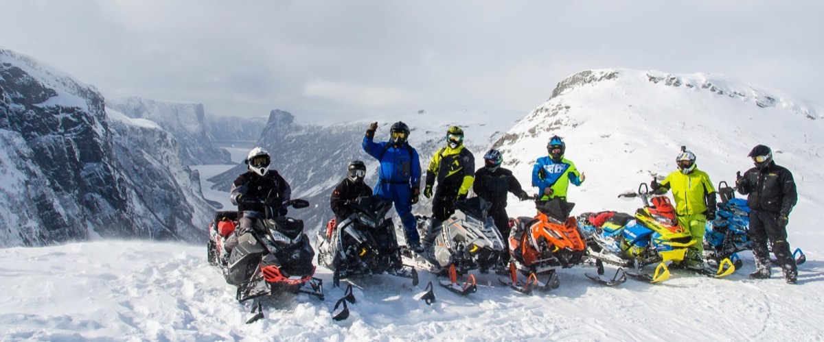 A group of riders and sleds on a snowy summit overlooking a long fjord and steep mountain walls
