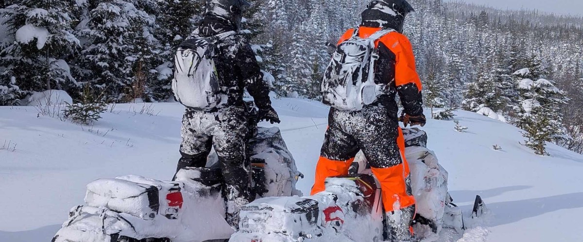 Two riders in snow-covered gear stand with their sleds in the trees